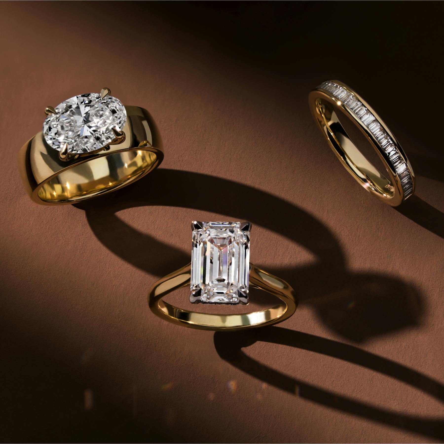 Three gold engagement and wedding rings set with diamonds, styled on a warm brown background with dramatic shadows.