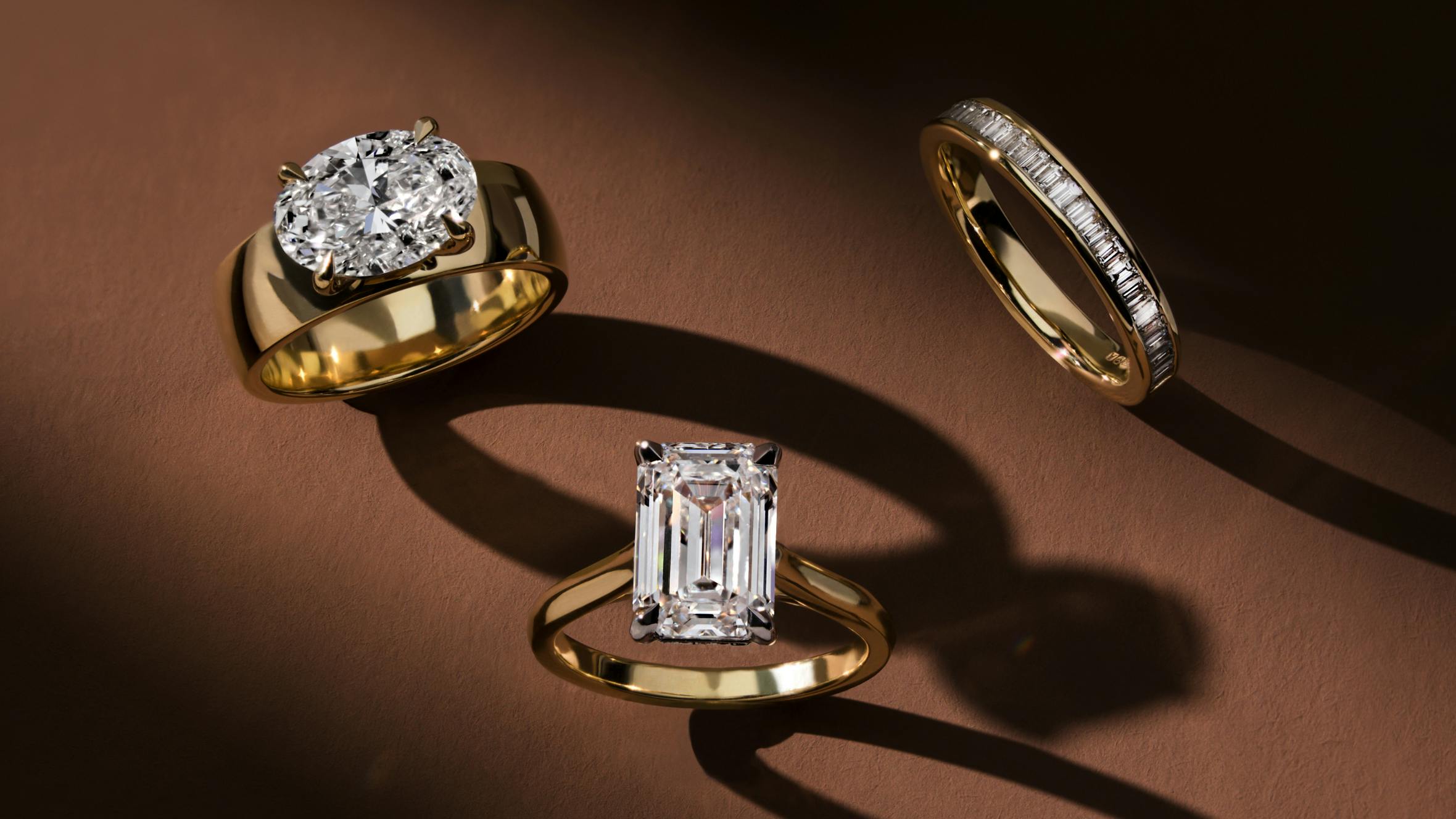 Three gold engagement and wedding rings set with diamonds, styled on a warm brown background with dramatic shadows.