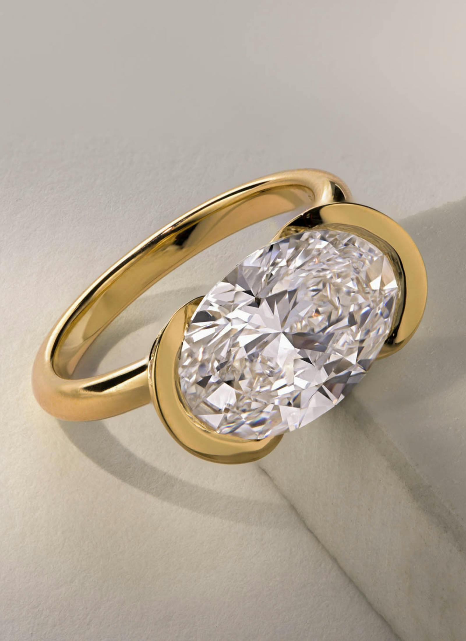 Unique bespoke engagement ring featuring an oval cut diamond in a partial rubover setting, crafted in yellow gold with an open, modern silhouette.
