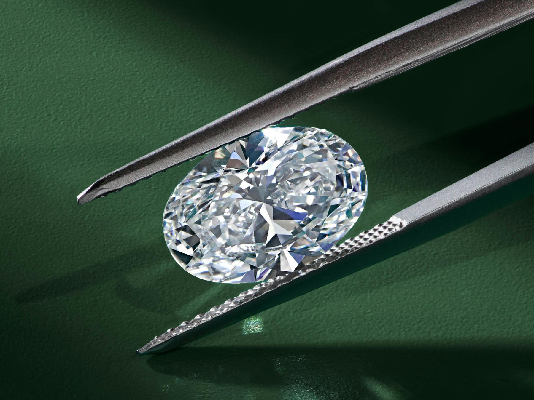 Close-up of a loose lab grown oval diamond held in precision tweezers against a dark green background, highlighting the stone’s brilliance and cut detail – showcasing lab grown quality.