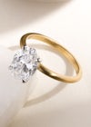 Solitaire yellow gold engagement ring with an oval diamond, with the base of the oval diamond resting on a tile, on a plain background with soft shadows