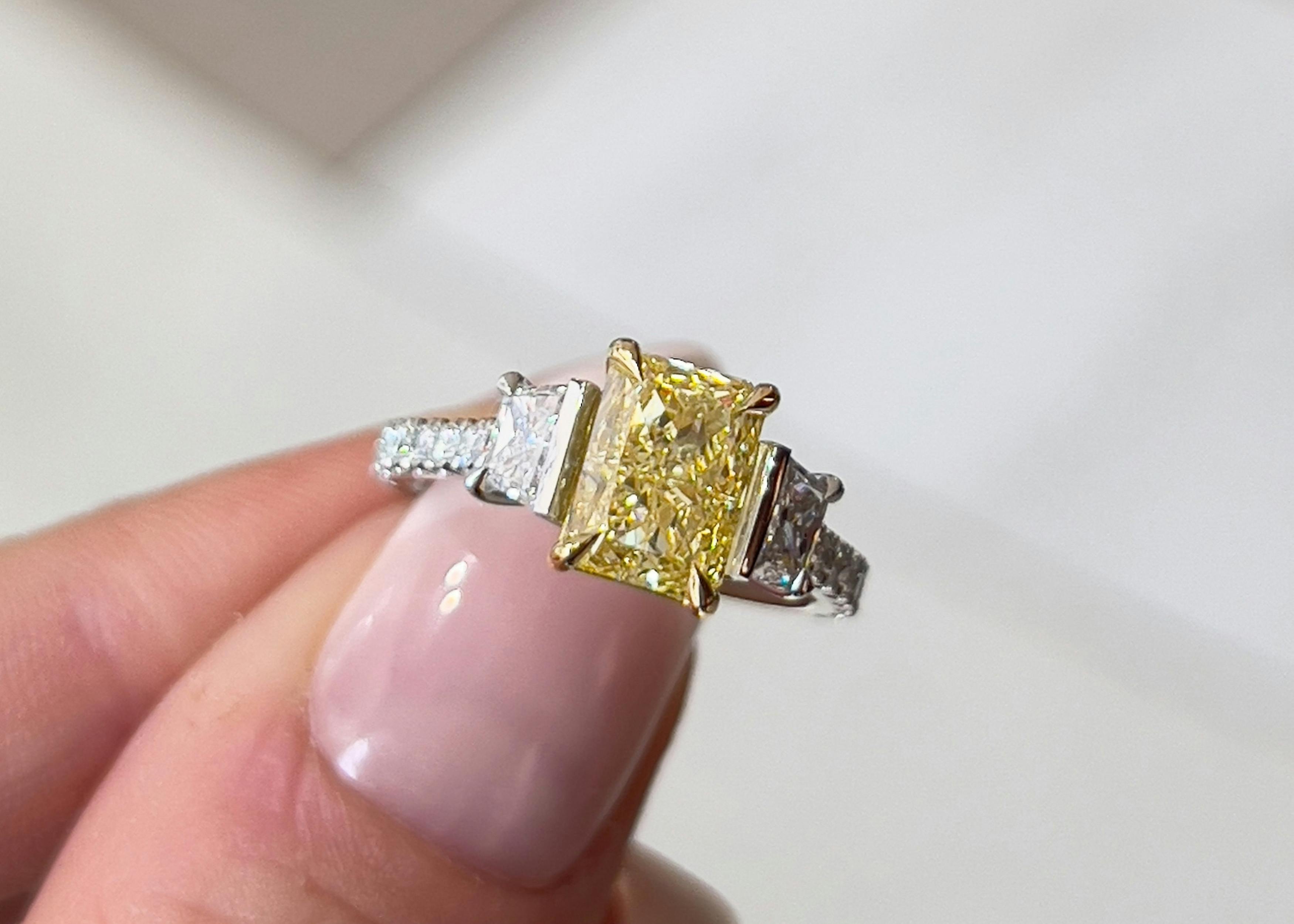 A close-up, aerial-view image of a manicured thumb and forefinger holding a 1.61ct radiant fancy yellow diamond yellow gold trilogy ring.