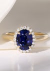 A deep blue, oval sapphire in a yellow gold halo ring setting, featuring tiny diamonds around the sapphire and a plain gold band