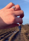 Woman showing off her custom engagement ring and humorously pretending to pick up her partner, who is standing further away from the camera