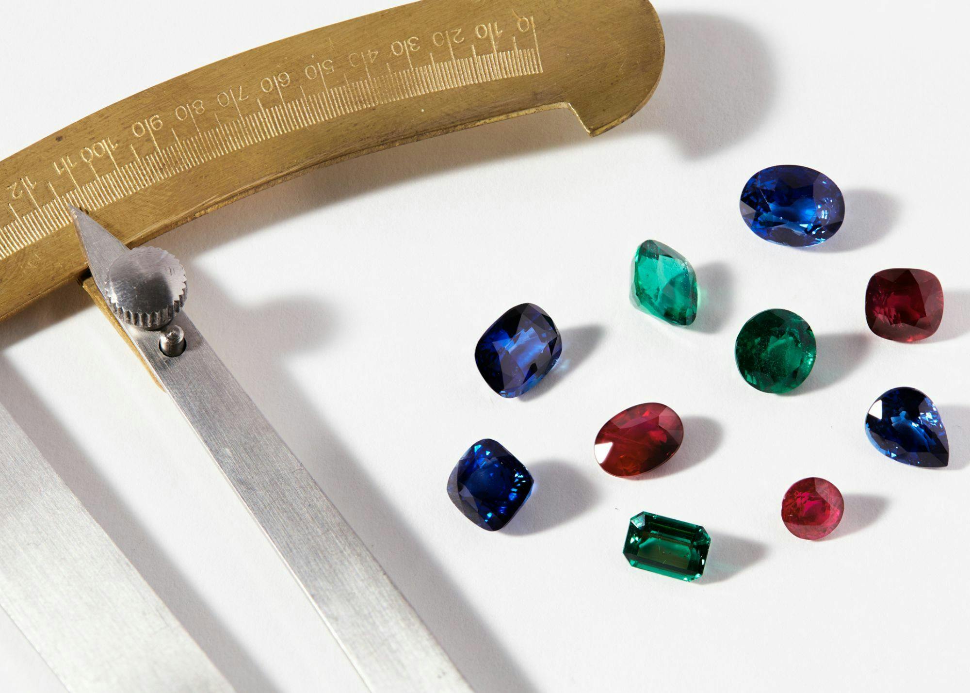 An aerial-view shot of scattered coloured gemstones, including rubies, emeralds, and sapphires, framed by a specialist measuring device.