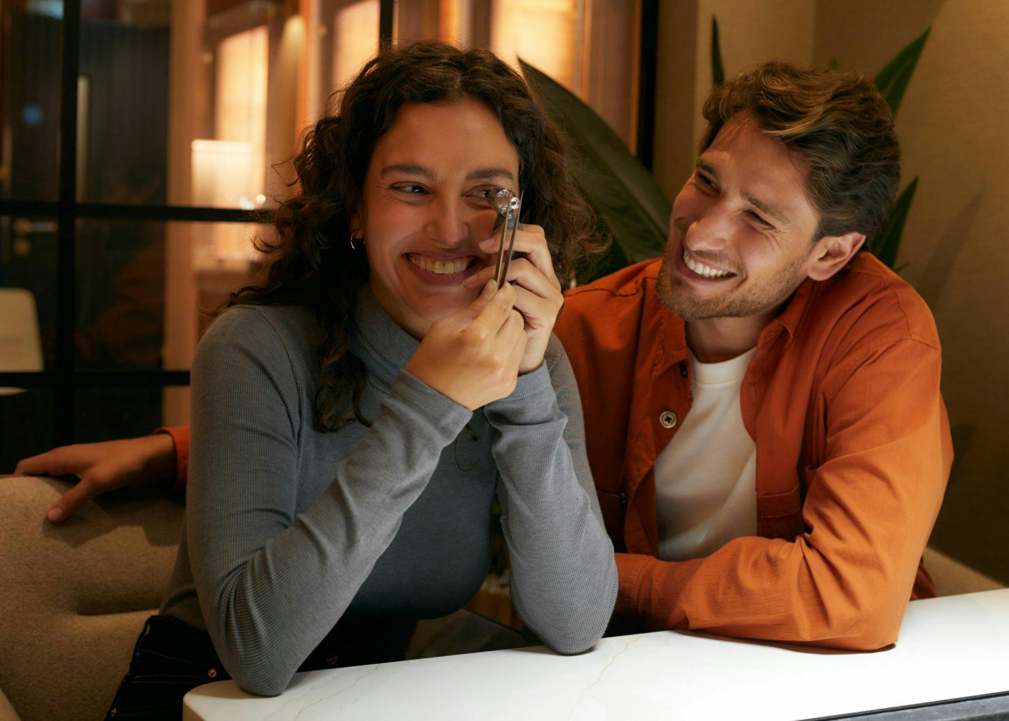 A smiling couple sit side by side. A man looks lovingly towards his girlfriend as she inspects a diamond using a loupe.