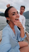 Woman shows off her bespoke sapphire engagement ring with her partner smiles and makes a shocked face behind her