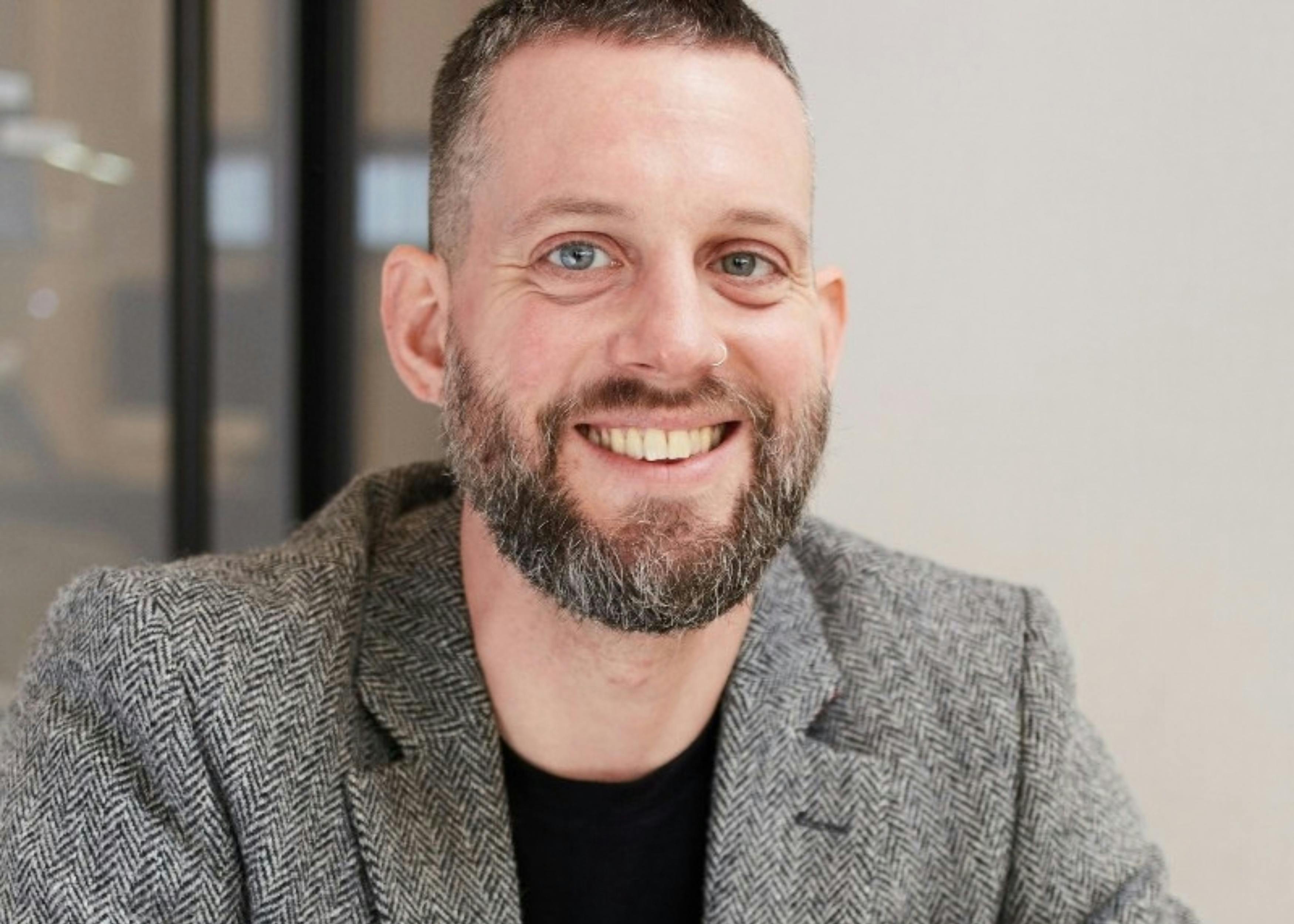 A front-view image of Eddi Norris, Queensmith's Sales Lead and Head of Gemmology. He is wearing a grey chequered suit jacket with a black t-shirt, smiling into the camera.