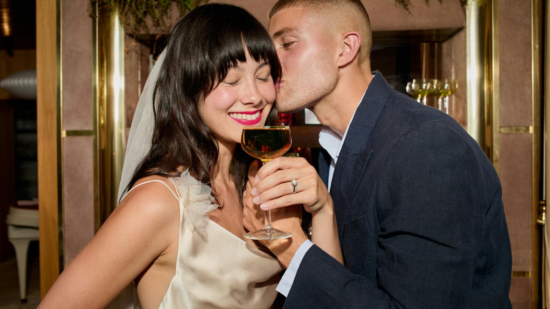 Bride smiles wearing an engagement ring and wedding ring, holding a glass of champagne as the groom kisses her cheek.