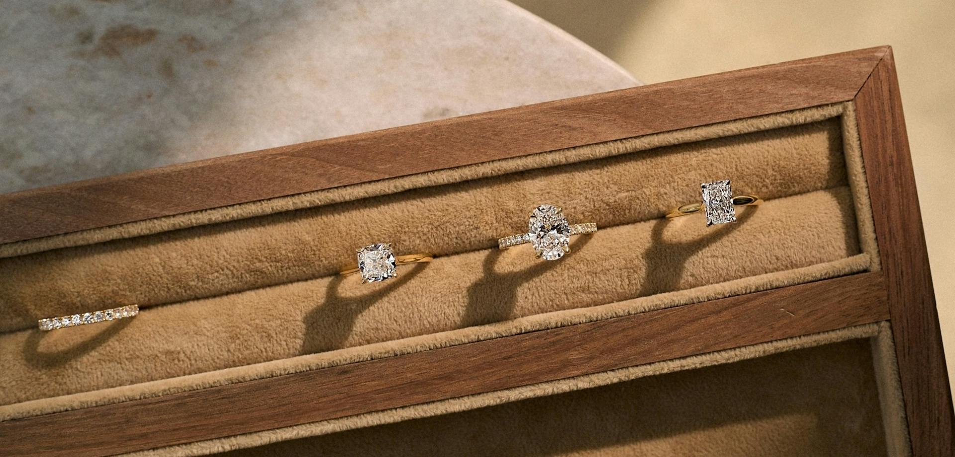 A close-up, aerial-view shot of three diamond engagement rings and one wedding band. They are set within a light leather viewing case.