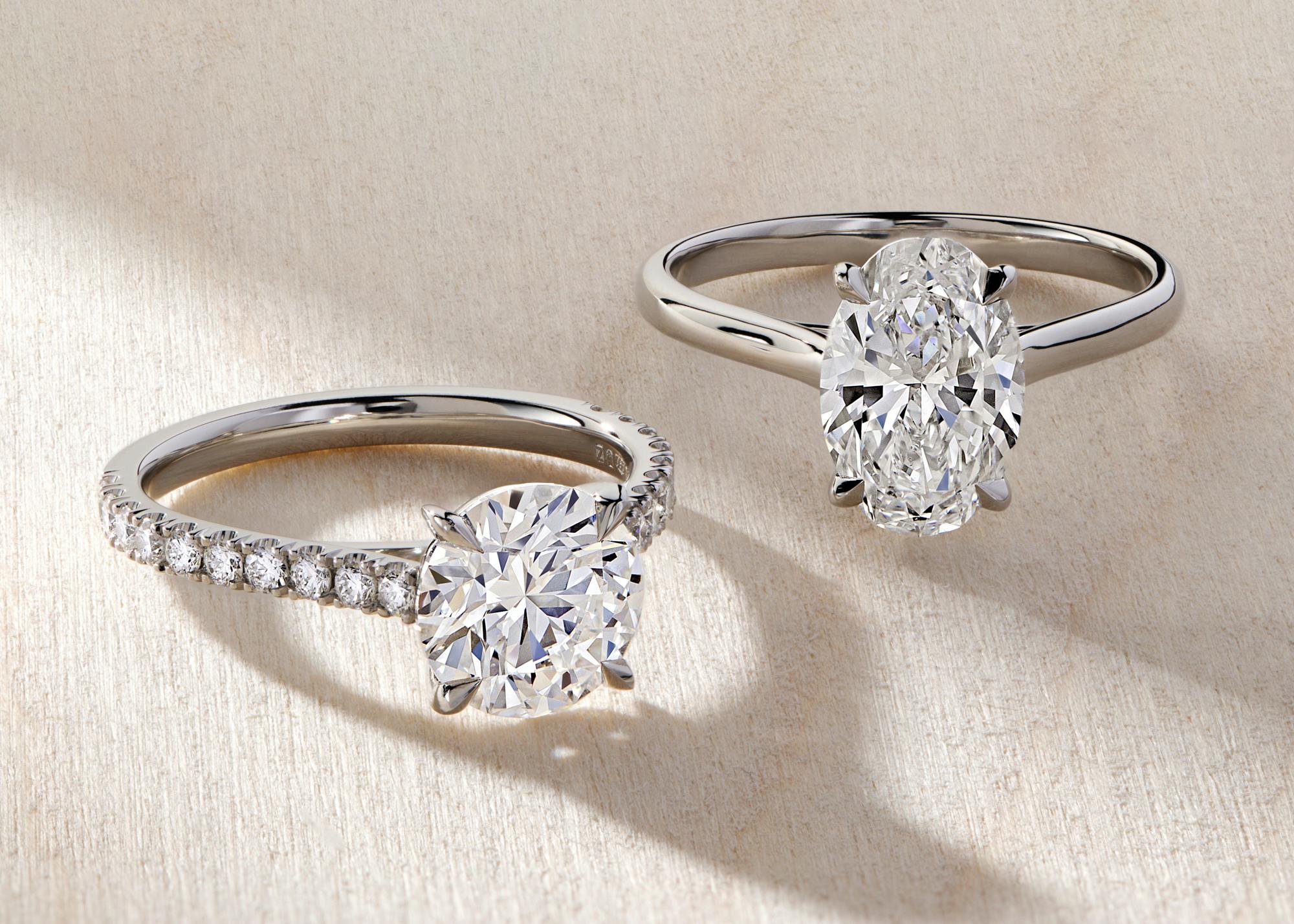 Two beautiful platinum engagement rings in a stream of sunlight on a beige background. One features a round diamond and scallop set diamond band, the other is a large oval diamond solitaire ring.