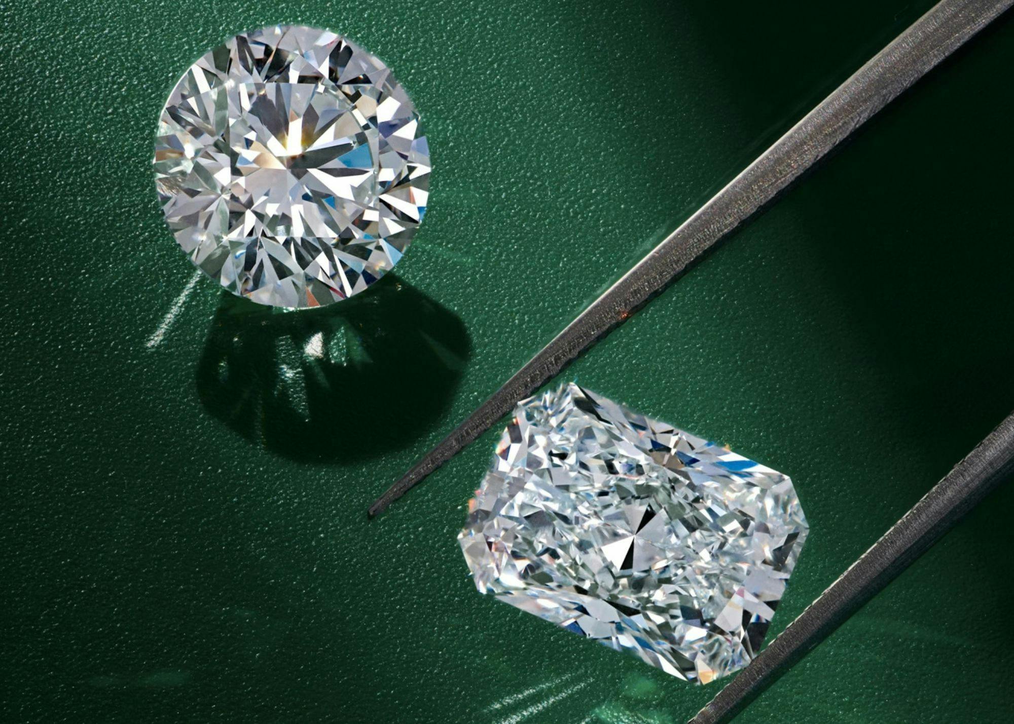 A close-up image of two loose diamonds with one between the prongs of a tweezer. Both diamonds are set carefully on a green textured surface.