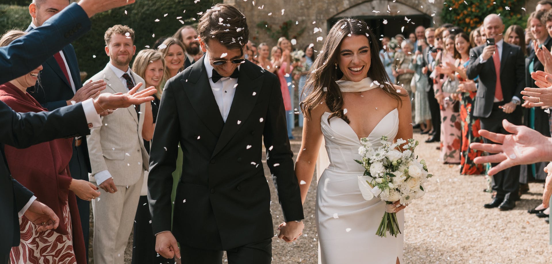 Bride and groom hold hands as family and friends throw confetti over them.
