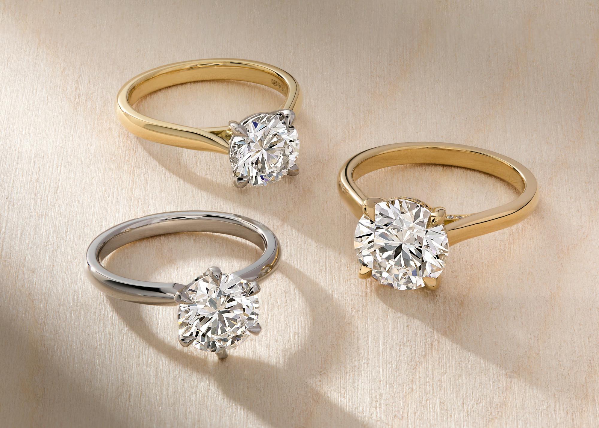 Three simple, round diamond engagement rings arranged on a beige background, two in yellow gold and one in platinum, with warm sunlight lighting them up from the right-hand side