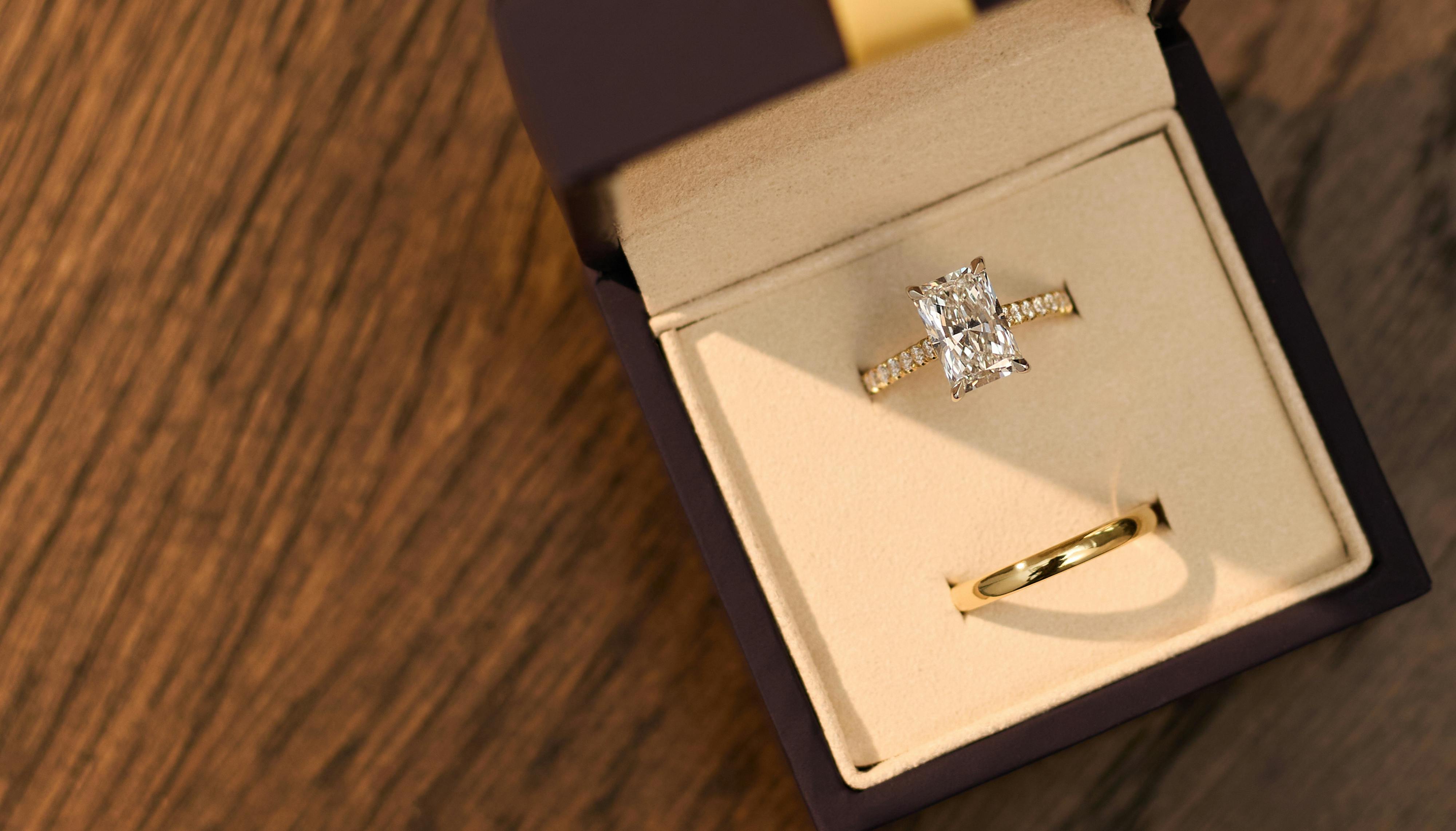 Aerial view of a radiant cut, yellow gold engagement ring in a ring box with a plain gold wedding ring next to it, the ring box sitting on a warmly lit wooden table