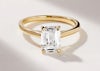 A big emerald cut diamond yellow gold solitaire engagement ring, on a plain white background with dramatic shadows either side of the beautiful gold engagement ring