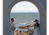 Bride and groom stand by large window overlooking the sea.