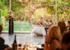 Woman stands with mic in front of bridge and groom and wedding guests giving a speech.