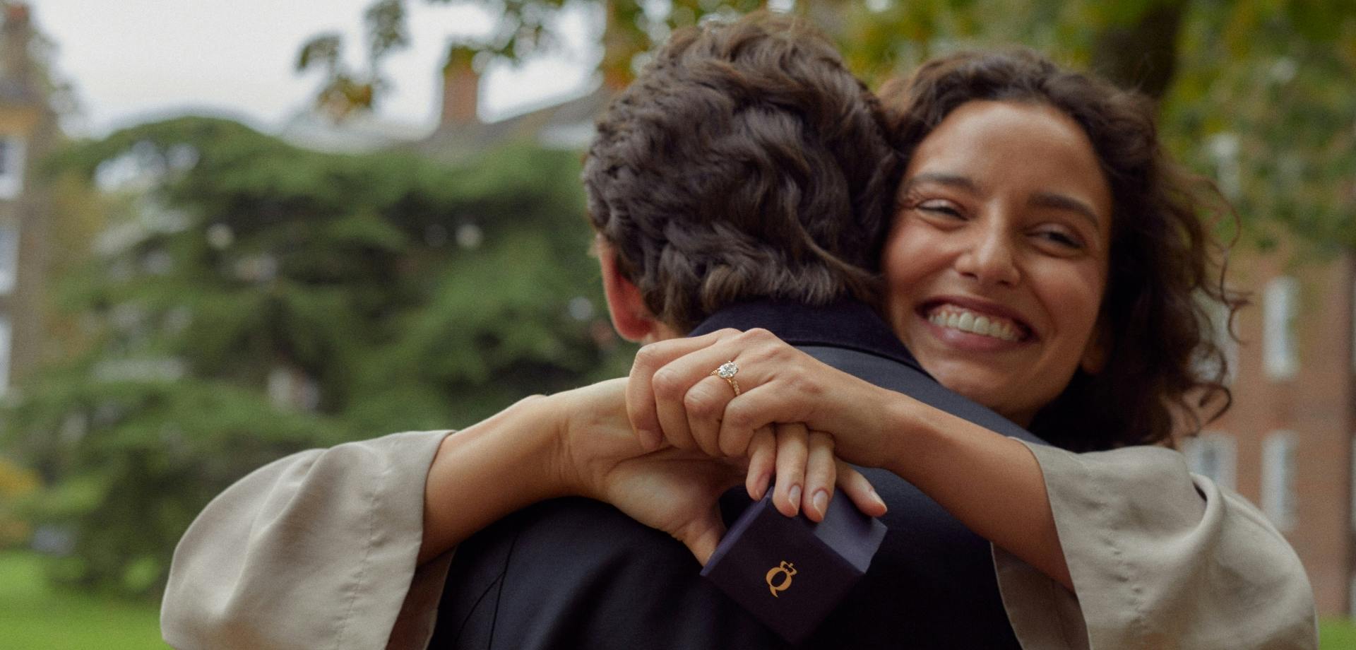 A close-up image of a couple hugging, taken from behind with the female's face visible and smiling. She is holding a Queensmith engagement ring box and her new diamond engagement ring is visible.