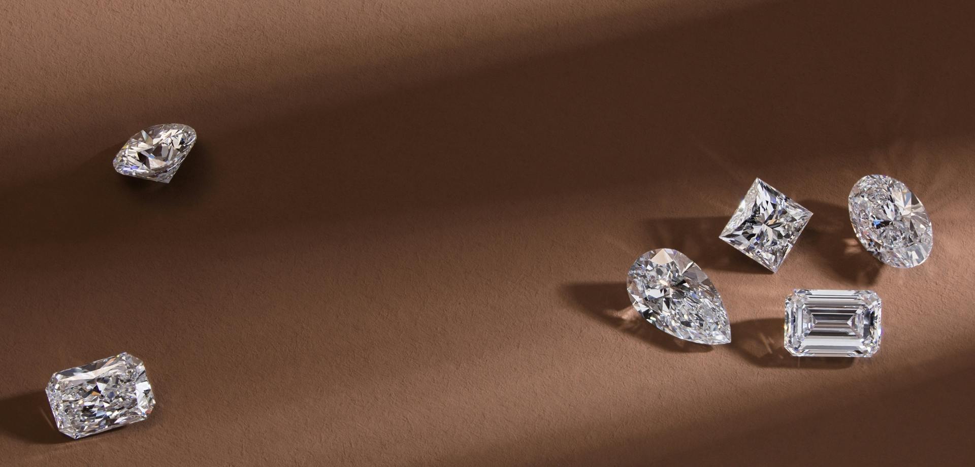 An aerial-view image of a variety of loose diamonds in cushion, marquise, oval, and emerald cuts on a dark brown, shadowed background.