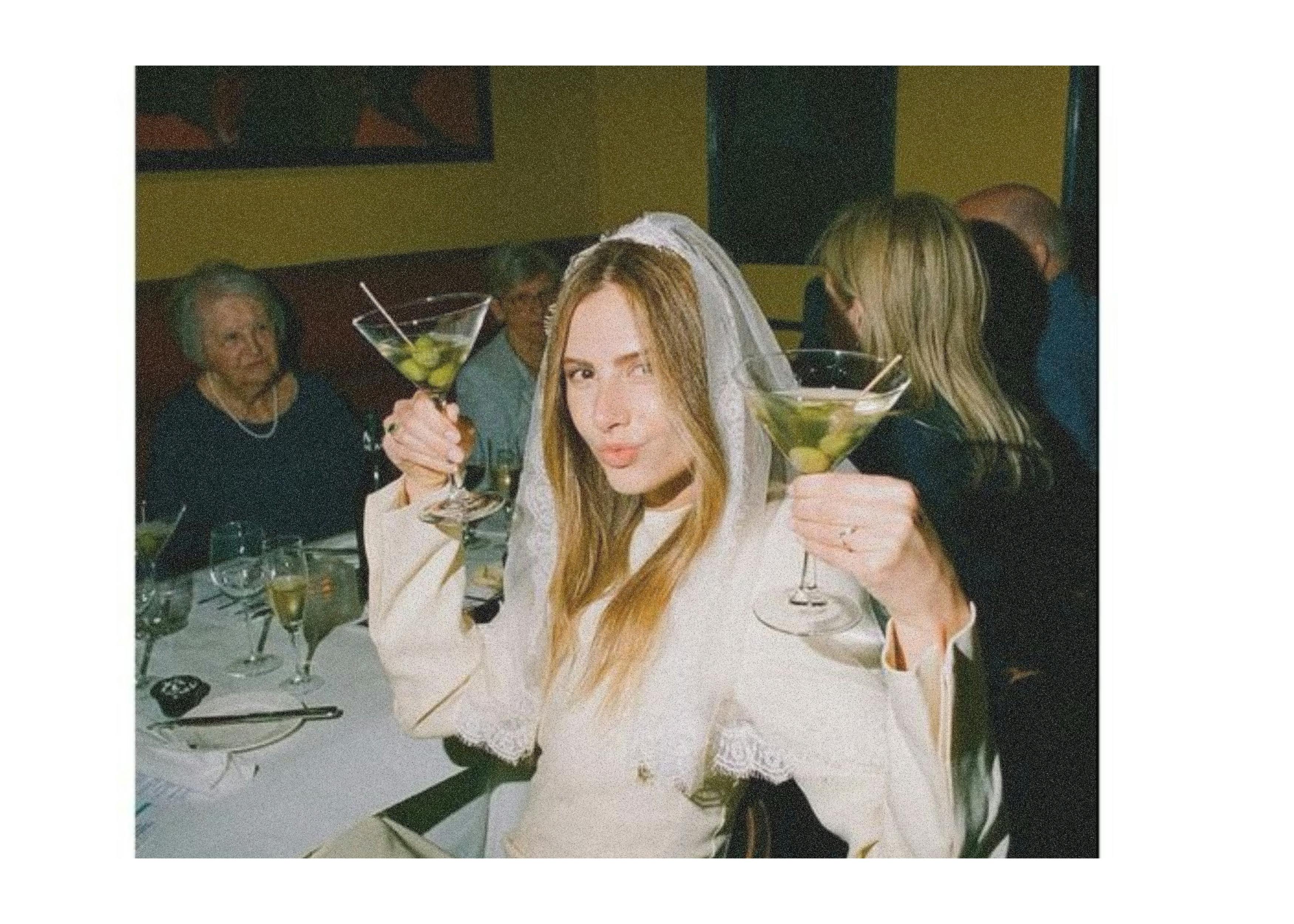 An image of a modern bride holding two full martini glasses while posing and pouting to the camera. She is sat at an informal dining table with her nearest and dearest.