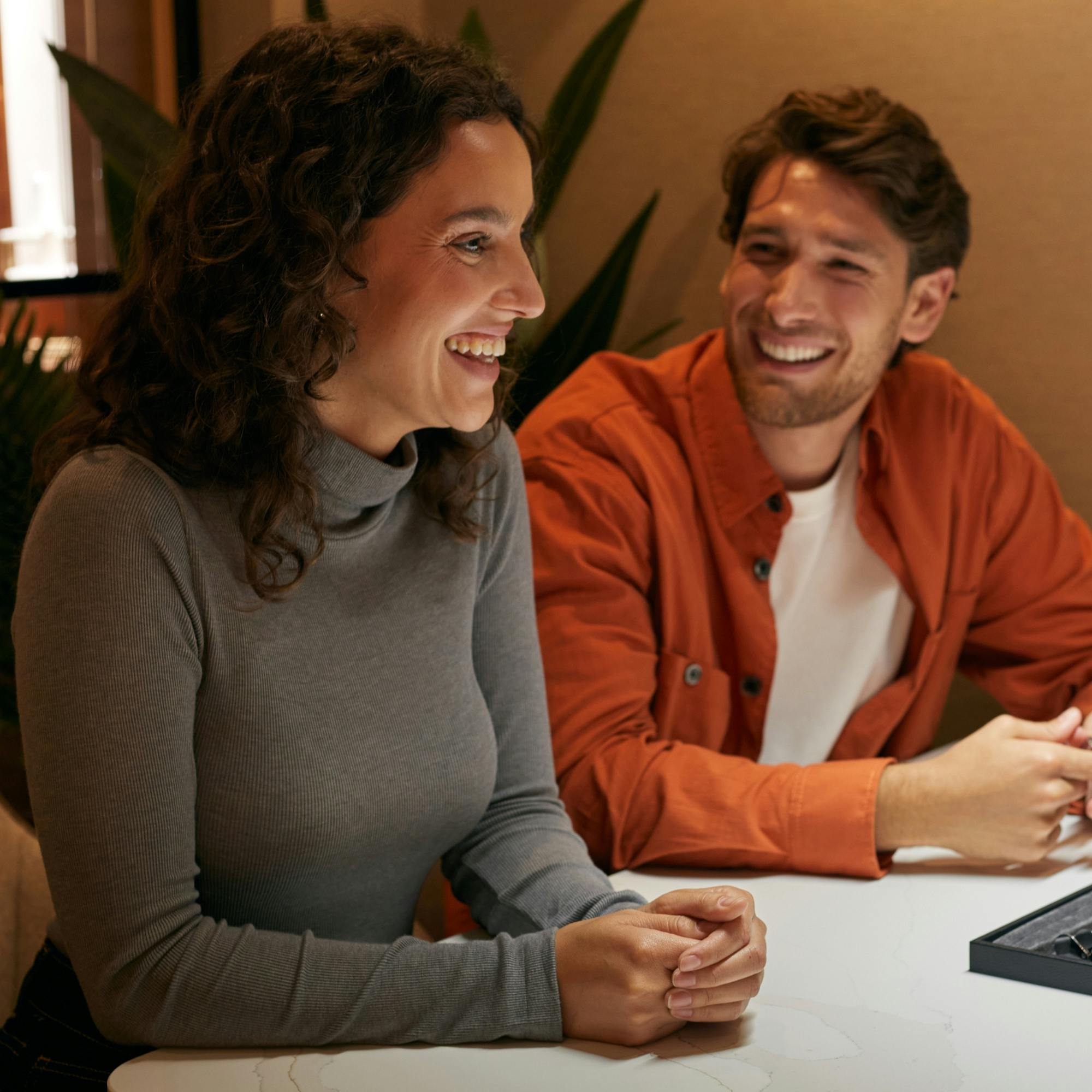A middle aged couple sit at a consultation desk smiling while designing their engagement ring.