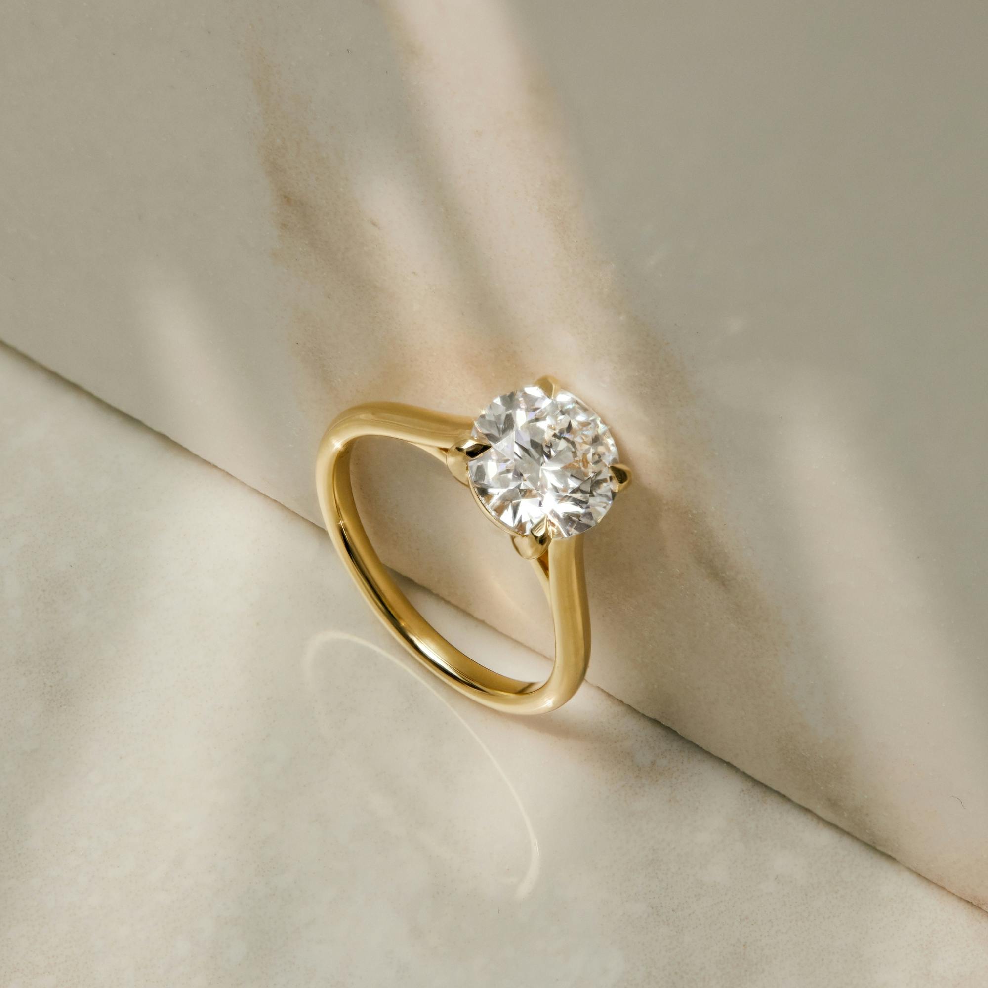 A side-view shot of a non-wedfit bespoke Temple solitaire in platinum, resting against a marble wall with sunlight striking its diamond.