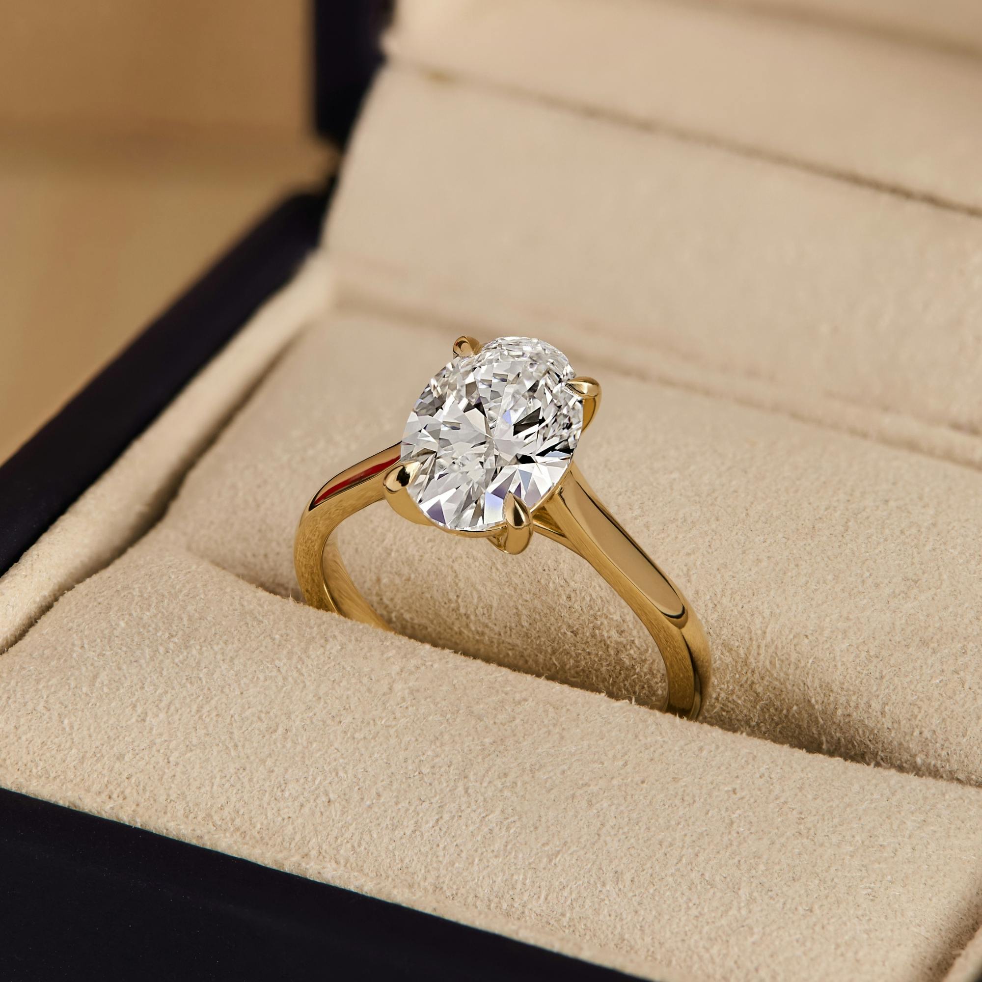 A close-up angled image of a oval diamond engagement ring in yellow gold sat in a neutral-coloured ring box.
