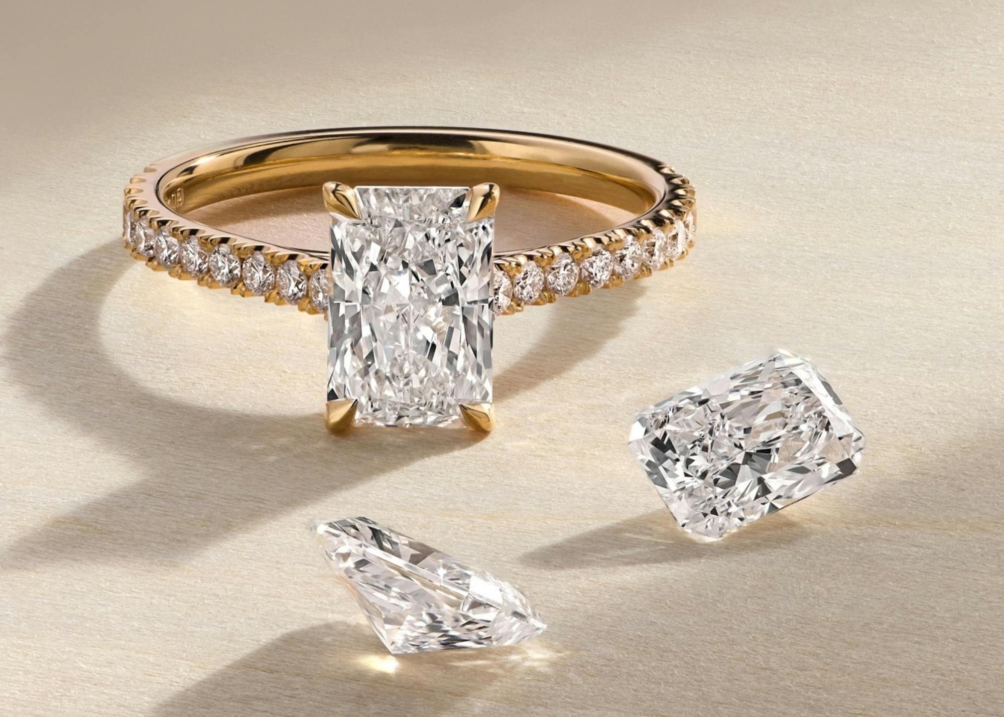 A close-up image of a radiant bardot diamond set within a scallop edged yellow gold engagement ring. Two loose diamonds are positioned in front of the ring.