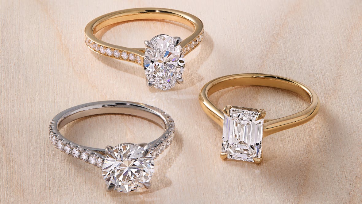 Three diamond engagement rings – two yellow gold rings, one with a pave diamond band and oval diamond, one with a plain band and emerald cut diamond. Third ring in platinum with scallop diamond band and large round cut diamond.