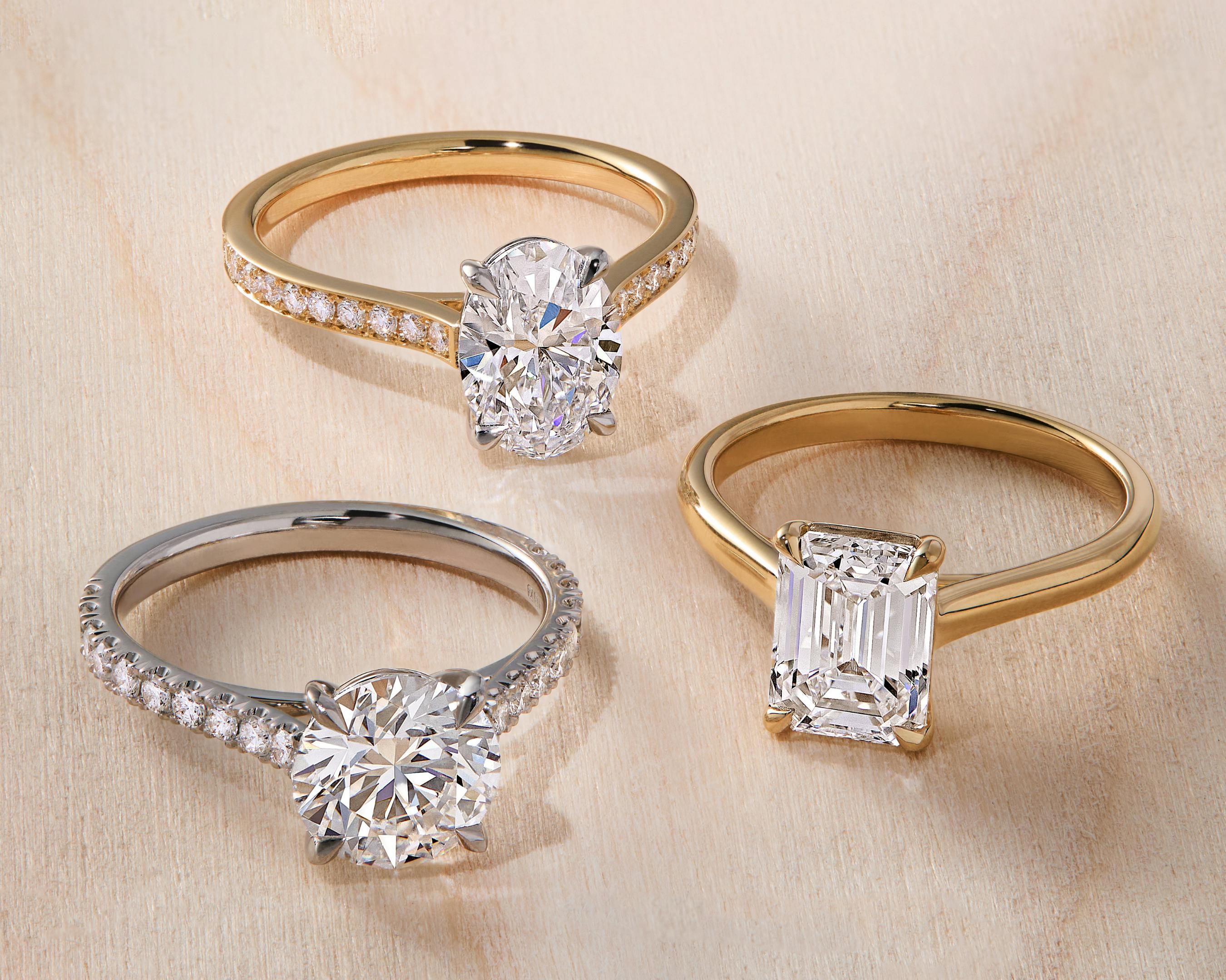 Three diamond engagement rings – two yellow gold rings, one with a pave diamond band and oval diamond, one with a plain band and emerald cut diamond. Third ring in platinum with scallop diamond band and large round cut diamond.