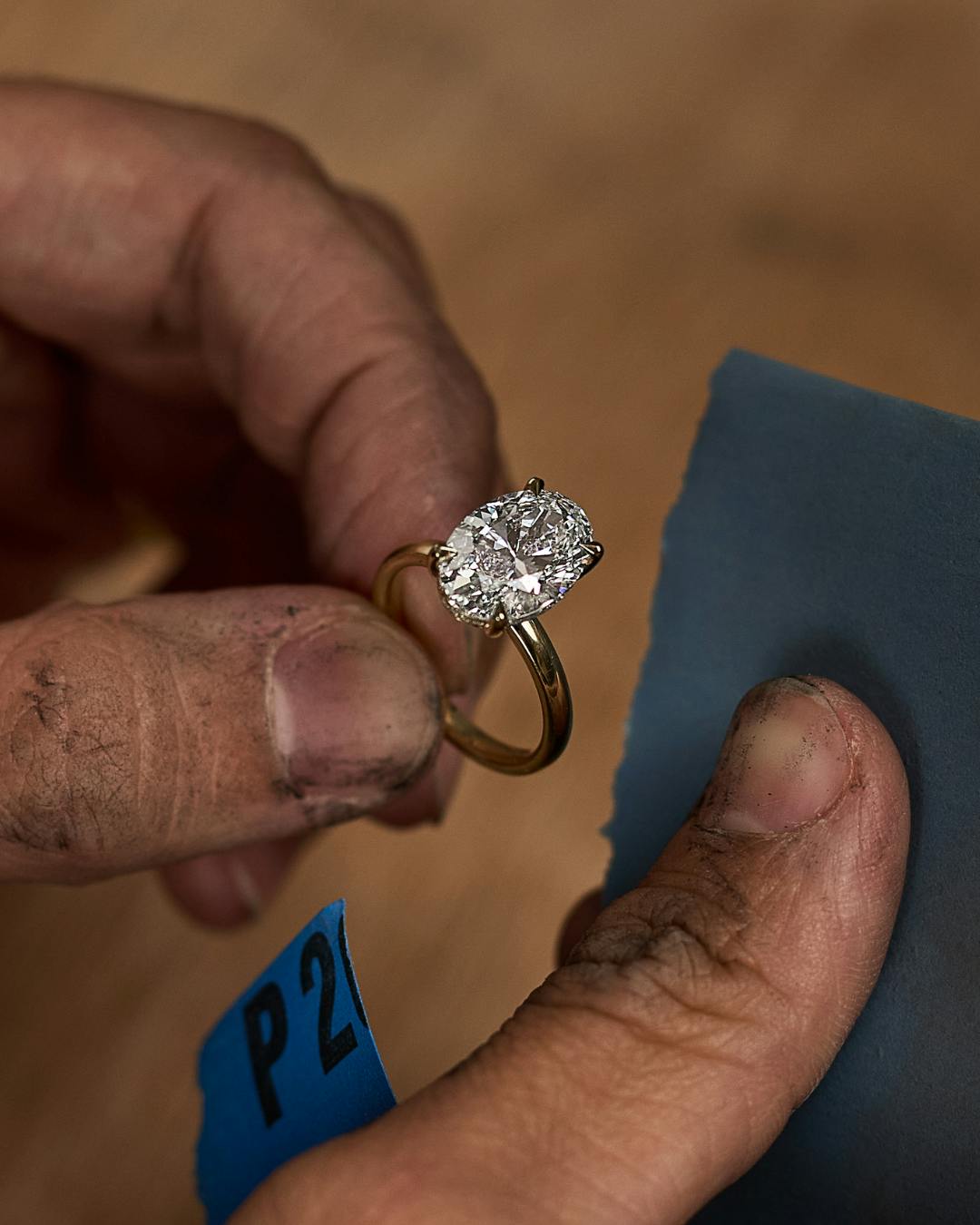 A close-up of a jeweller’s hands holding a yellow gold engagement ring with a large oval diamond, preparing to polish it using a blue polishing cloth. The hands show signs of craftsmanship.
