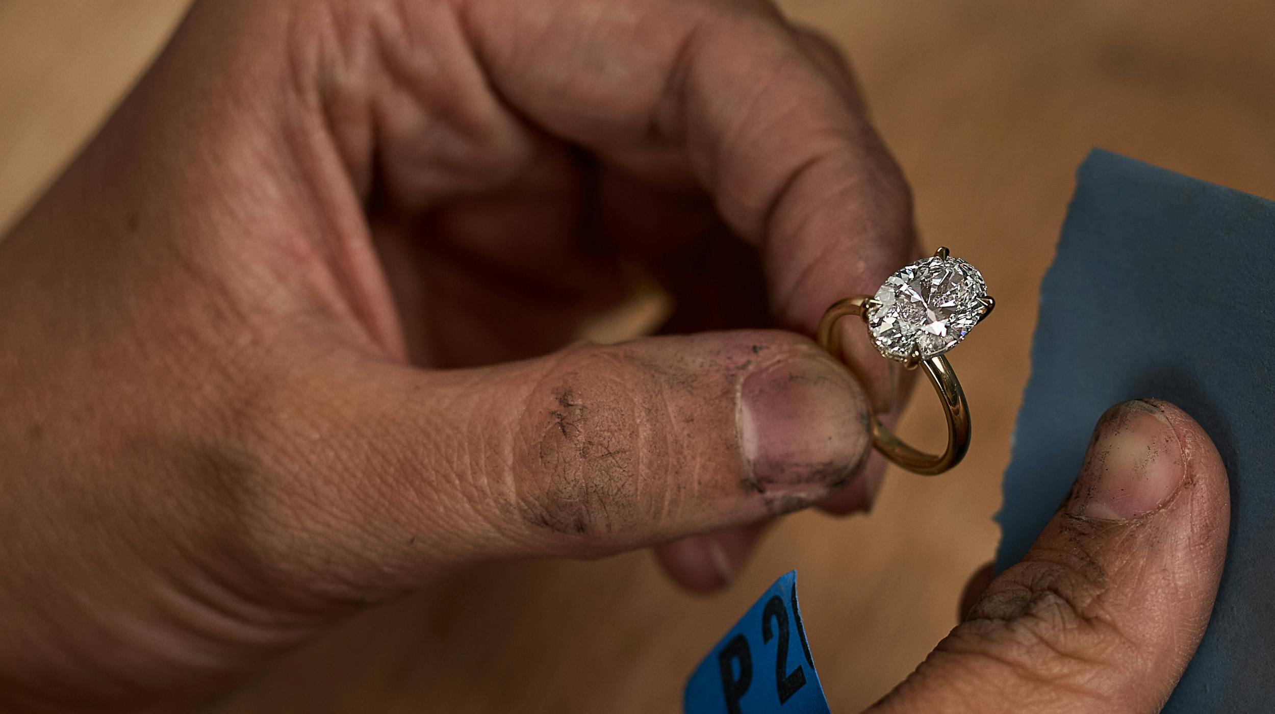 A wide shot of a jeweller carefully holding a yellow gold diamond engagement ring between their fingers, about to polish the metal band using a blue cloth. The diamond sparkles under the light, while the jeweller's hands show evidence of skilled, hands-on work.