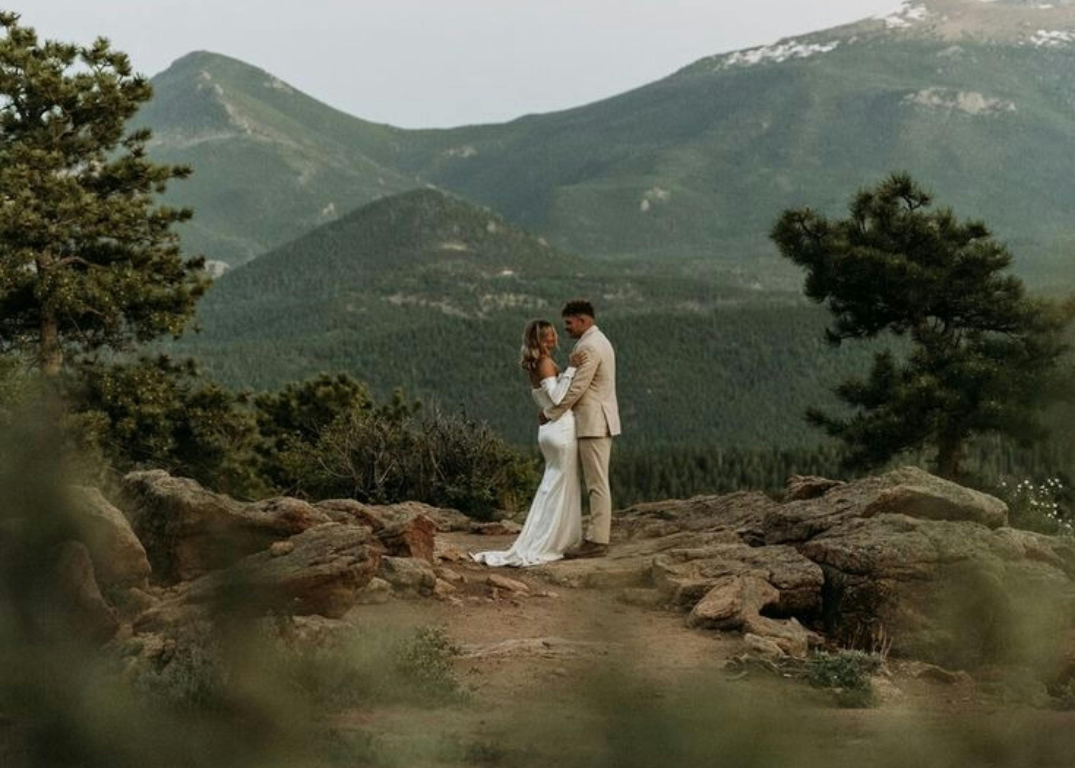 An image of a newly married couple embracing and looking at each other, positioned in the centre of the frame. They are positioned in front of a grand mountain background, framed by greenery.