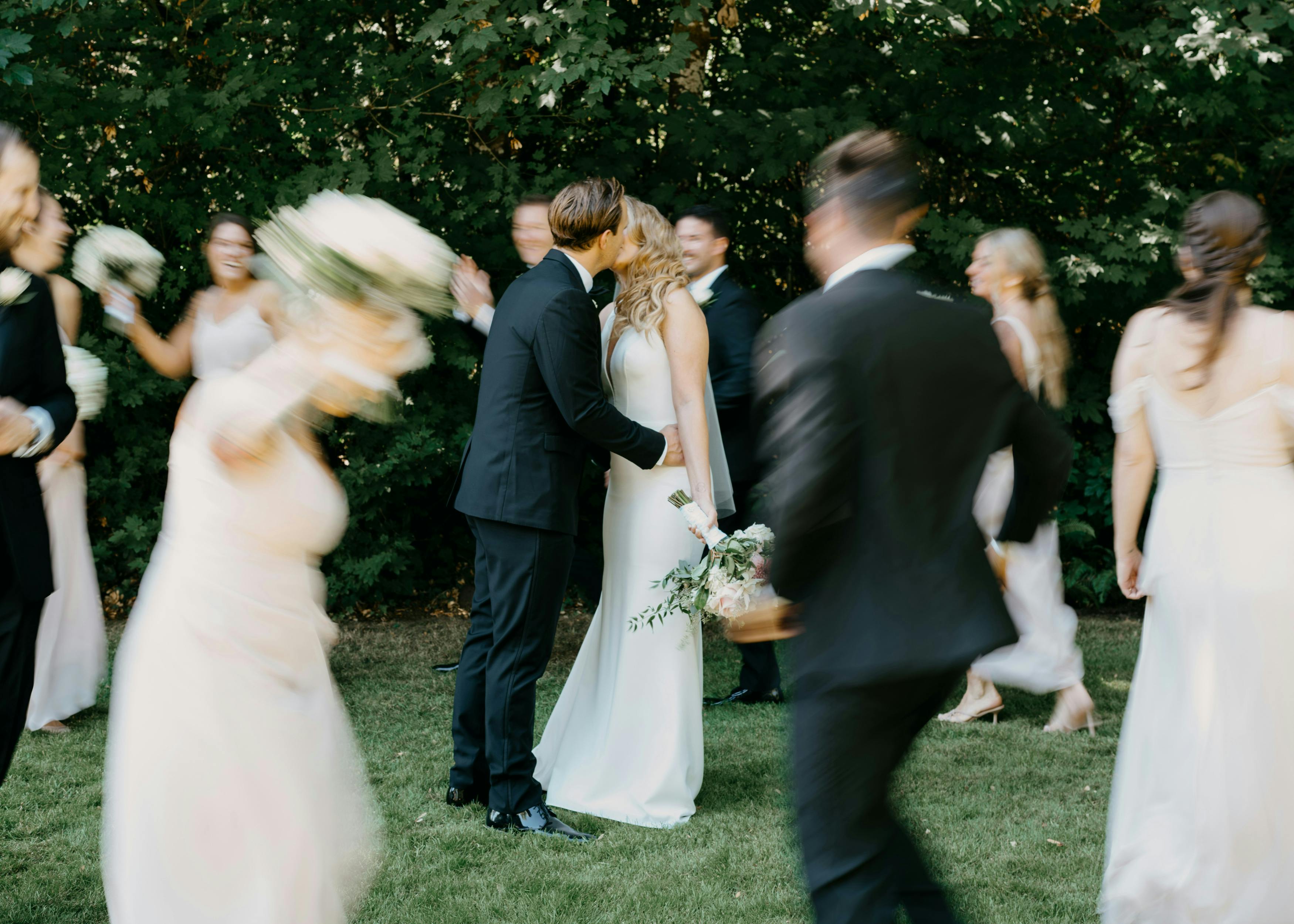 Newly married couple kissing outdoors as wedding guests move joyfully around them, creating a sense of motion and celebration.